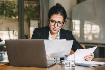 Photo of adult businesswoman 30s wearing formal clothing and eyeglasses working in office on laptop and examining paper documents, while holding in hand