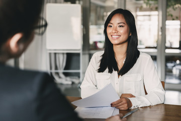 Portrait of joyful asian woman smiling and holding resume, while sitting in front of businesswoman...