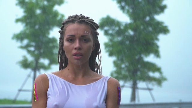 A sad attractive girl is standing and crying in the rain in a wet white dress.