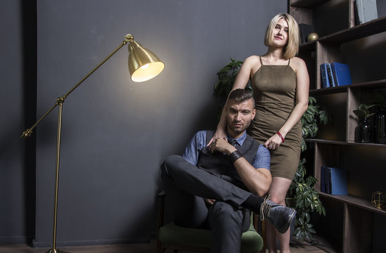 woman near with successful rich man. Young successful businessman in chair and his sexy woman in dress in luxurious apartment of wealthy house. Portrait of beautiful lovers in the interior