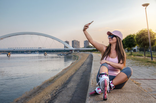 Attractive young adult woman taking selfie after roller skating by the river at sunset 