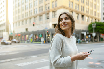 Beautiful young woman on the boulevard in urban scenery, downtown, at sunset, holding smartphone