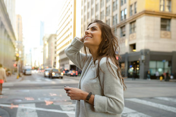 Beautiful young woman on the boulevard in urban scenery, downtown, at sunset, holding smartphone