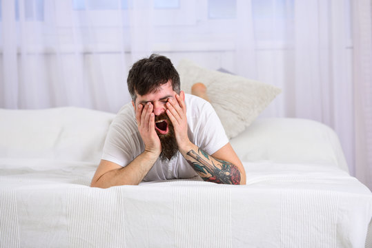 Guy on sleepy yawning face laying on edge of bed on white sheets. Bedroom concept. Man laying on bed, white curtains on background. Macho with beard and mustache relaxing, having rest.