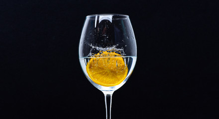Wineglass filling with water with splashes on black background. Refreshing drink concept. Glass with water and slice of citrus pouring with liquid with splashes and drops of water