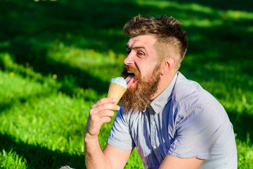 Man with long beard eats ice cream, while sits on grass. Bearded man with ice cream cone. Man with beard and mustache on happy face licks ice cream, grass on background, defocused. Delicacy concept.