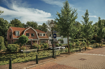 Tree-lined canal with aquatic plants, boats on the banks and brick houses on a sunny day in Weesp. Quiet and pleasant village full of canals and green near Amsterdam. Northern Netherlands.