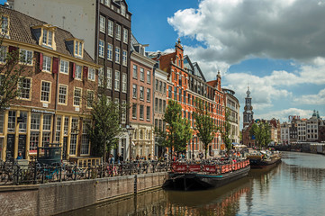 Fototapeta na wymiar Wide canal with typical houses, steeple, moored boats and blue sky in Amsterdam. Famous for its huge cultural activity, graceful canals and bridges. Northern Netherlands.