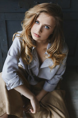 Fototapeta premium Fashionable and beautiful blonde model girl with light makeup and amazing blue eyes in unbuttoned blue shirt and in khaki pants sits and posing in art interior
