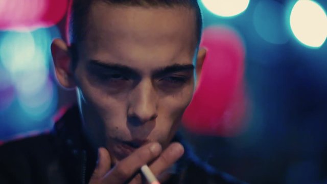 Handsome young man with a hipster ponytail smokes cigarette outside the bar, club at night. City lights on the background. Brutality, man&rsquo;s style, criminal. Male portrait