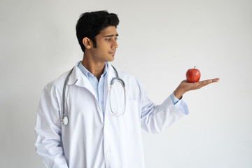 Pensive handsome Indian male doctor holding apple on palm and looking at it. Serious professional...