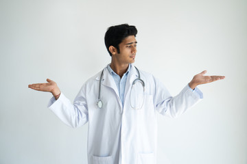 Pensive handsome Indian male doctor holding out hands while comparing products. Introspective young...