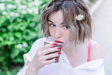 Attractive girl with red lips and manicure whith flower in her hair is sitting in the park and drinking coffee