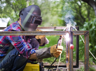 Man in a protective mask welding metal construction outside the house on a summer day