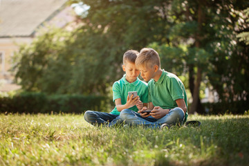 Two kids brothers playing games on smartphone with excitement while sitting on grass in park.
