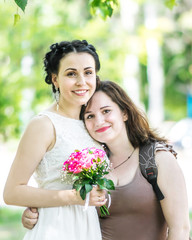 Portrait of two young pretty women hugging each other in green summer park. Pretty females bride with bouquet of roses and her friend or sister smiling and looking into camera