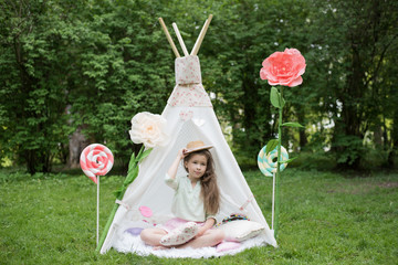 Little girl lying and playing in a tent, children's house wigwam in park. Happy mothers day. Child with big donuts and heart-shaped pillows. Summer, childhood concept. © MartaKlos