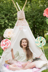 Little girl lying and playing in a tent, children's house wigwam in park. Happy mothers day. Child with big donuts and heart-shaped pillows. Summer, childhood concept. © MartaKlos