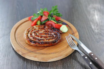 Sausage is spiral and cooked on the grill. Next slices of fresh vegetables. In the frame cutlery. Dark background. Close-up.