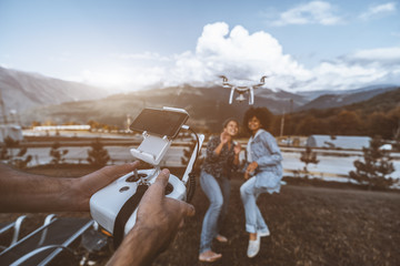 Wide-angle shot of the operator's hands with remote controller in the foreground driving the flying drone in front which filming two girls of different races posing for the broadcast, mountains behind