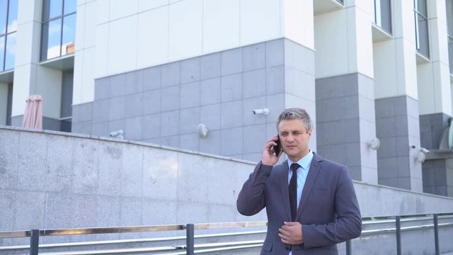Attractive middle-aged man. Serious businessman Talking on the phone An image of a general plan, against the background of modern office buildings. A solid man in a jacket and tie 4K