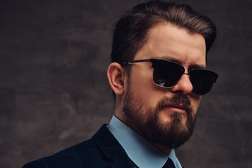 Close-up portrait of a handsome fashionable middle-aged man with beard and hairstyle dressed in an elegant formal suit and sunglasses. Isolated on a textured dark background in studio.