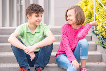 Happy teenage boy and girl smiling outdoors. Young sister and brother teens sitting on the stairs and look at each other.