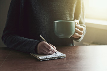 female hands with pen writing on notebook with morning coffee or tea