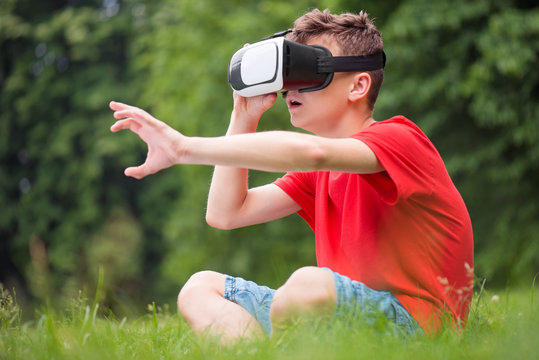 Playful teen boy using virtual reality goggles outdoor in summer park. Teenager looking in VR glasses. Child have fun or shock - experiencing 3D gadget technology.