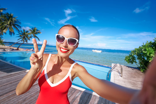 Vacation and technology. Colorful portrait of pretty young woman taking selfie portrait near swimming pool on tropical beach.