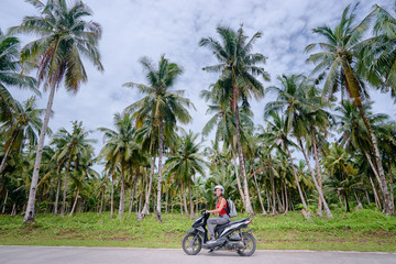 Naklejka premium Podróże i transport w tropikach. Młoda piękna kobieta w kasku, jazda na skuterze na drodze z palmami.