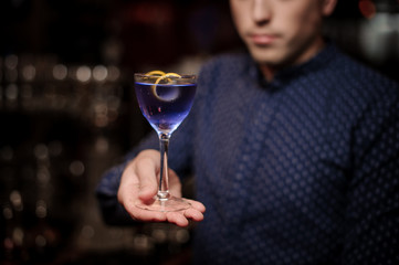 Bartender holding a glass of violet cocktail decorated with an orange pill