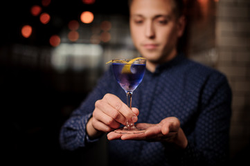 Young barman holding a glass of violet cocktail decorated with an orange pill