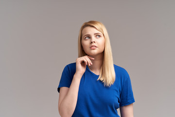 A pensive blonde woman, being deep in thought, holds her hand under her chin as she tries to...