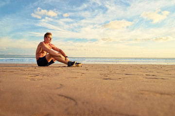 Fototapeta na wymiar Relaxation after sports. Young muscular man sitting on the sea beach enjoying sunset.