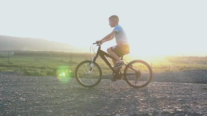 Little kid riding bike in summer top of mountain at beautiful sunset sky background. Beautiful caucasian small boy of 8 years riding on bicycle in mountainous area over river on sunrise or sunset