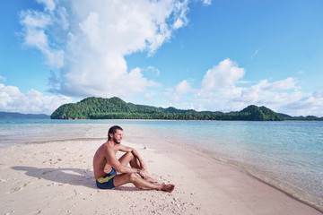 Fototapeta na wymiar Tropical vacation. Relaxed young man sitting on the sand beach enjoying the island view.