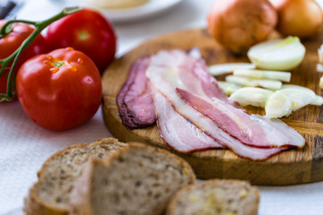Raw bacon on cutting desk with onion and garlic and tomatoe behind.