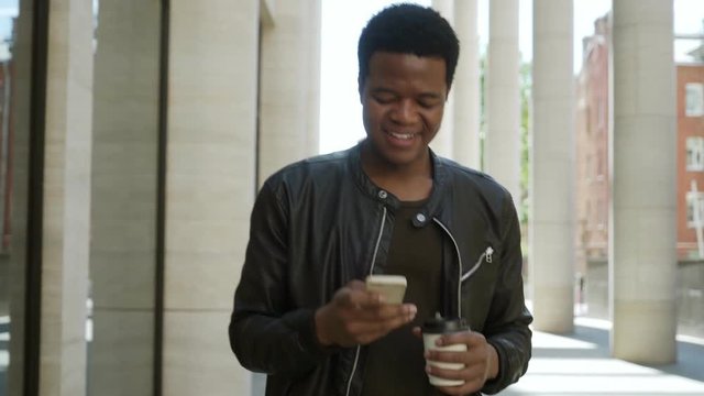 Follow shot of young African man in leather jacket walking down street with coffee in his hand, text messaging on phone and smiling