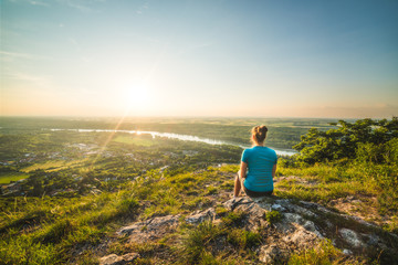 Fototapeta premium A Girl Sits on the Edge of the Cliff and Looking at the Sunset and the Valley with the River