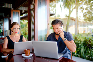 Coworking and freelance concept. Young  bearded man  and young woman working together on laptop computers while sitting on cafe terrace.