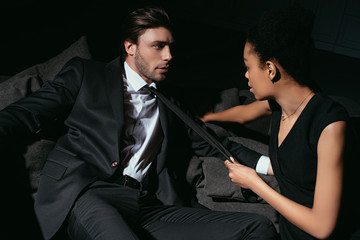 young multiracial sexy couple in black clothing on sofa on dark background