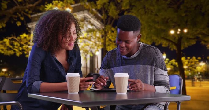 Smiling black male and female meeting for coffee use tablet computer, A millennial man and woman sit outside at night using touchscreen devices, 4k