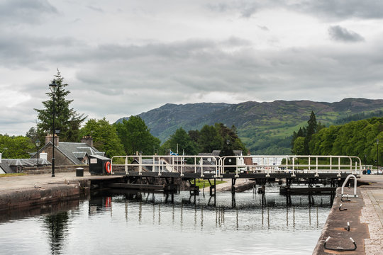Fort Augustus, Scotland - June 11, 2012: Lock with water flowing over closed dark brown doors on Oich River Canal showing green grass and white houses on both sides under heavy gray sky. Hills