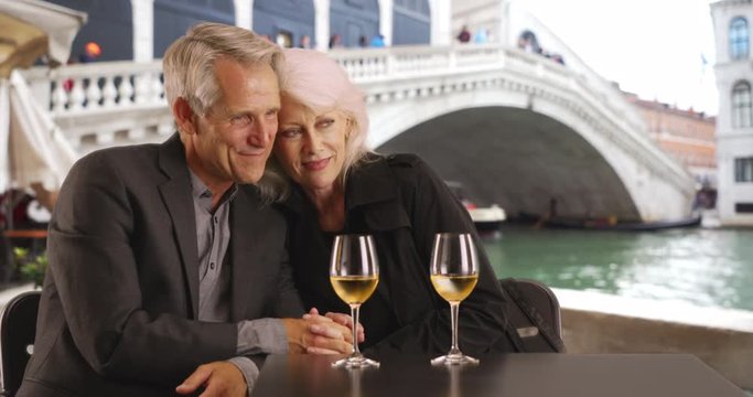 Lovely senior couple on holiday in Venice, Happy Caucasian male and female sit by the Grand Canal with a glass of wine, 4k
