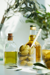 glass with spoon and green olives, jar, various bottles of aromatic olive oil with and branches on white table