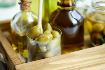 close up view of glass with spoon and green olives, jar, various bottles of aromatic olive oil on wooden tray