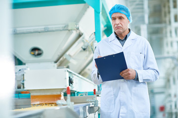 Portrait of senior factory worker doing  production quality inspection in food industry holding clipboard standing by conveyor belt and holding clipboard, copy space