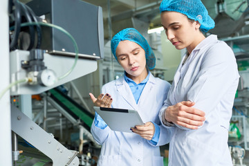 Portrait of two female specialists wearing lab coats working at modern food factory and checking production process via digital tablet standing by power units in workshop, copy space