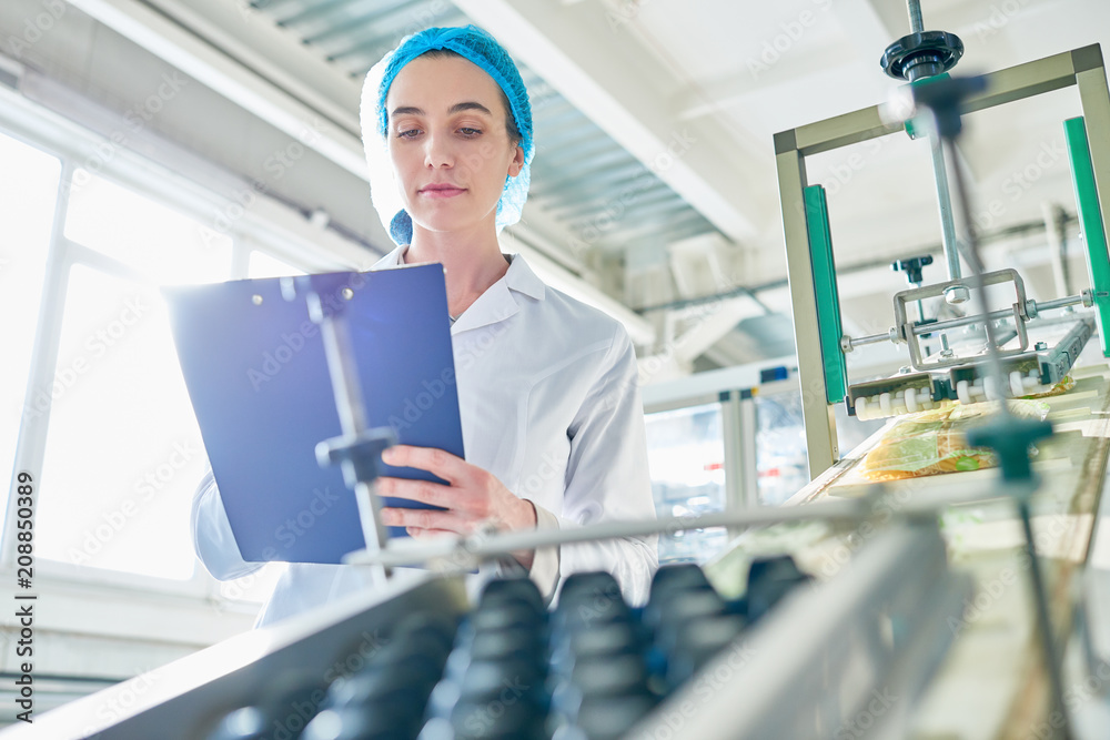 Wall mural Low angle portrait of  young female worker wearing lab coat standing by  power units and writing on clipboard  in clean production workshop, copy space